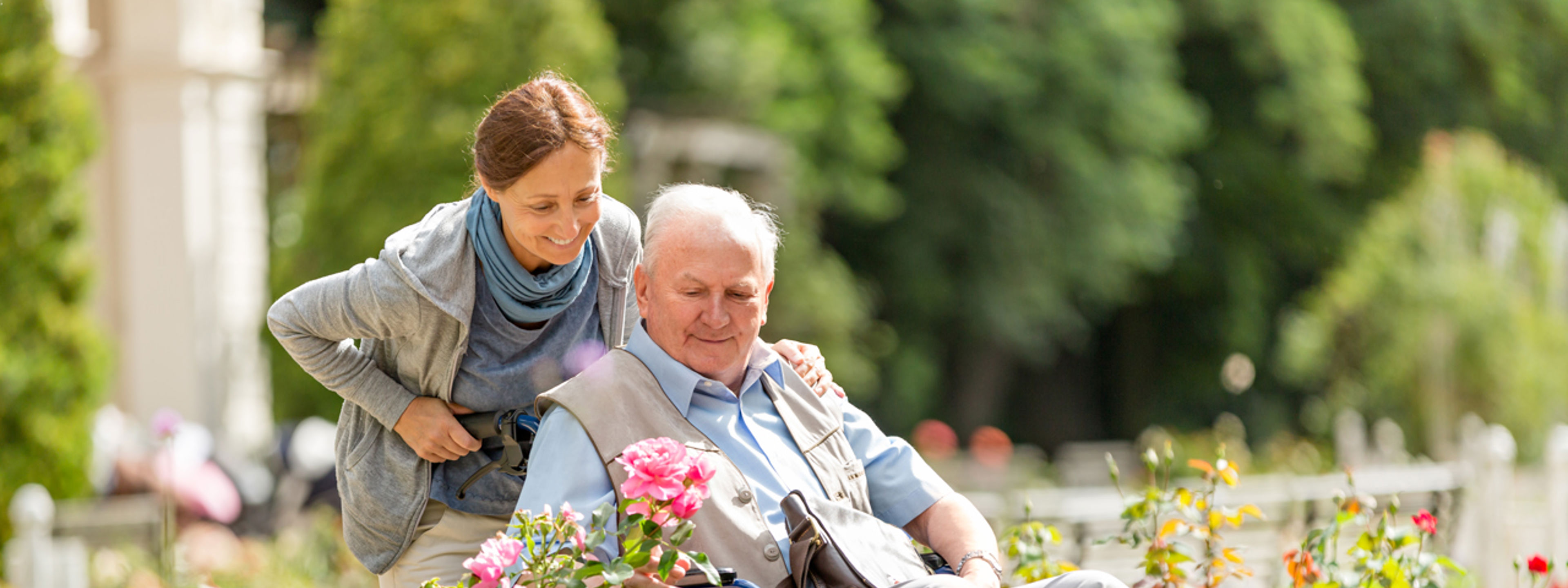 Ein eldre mann ser på blomar medan ei kvinne trillar han i rullestol.